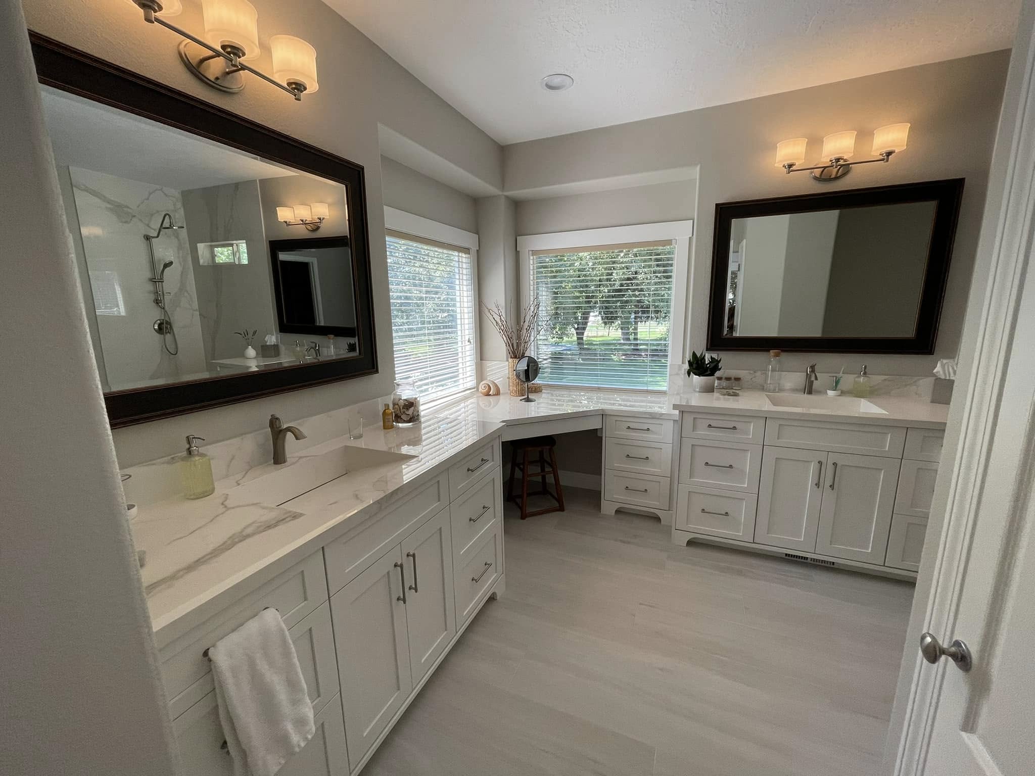 Finished master bath with wraparound white vanities, a dressing counter beneath corner windows, dark-framed mirrors and light tile flooring.